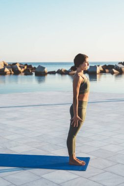 Yeşil spor kıyafetli zayıf bir kadın mavi hasırın üzerinde duruyor ve sabah su kenarında yogaya başlıyor.