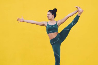 Spor kıyafetli yetişkin bir kadın, mindere basıp yoga yapıyor. El ele verip poz veriyor. Boş sarı duvarın arka planında tablet kullanarak video dersinden öğreniyor.