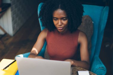 Afro-Amerikan kıvırcık saçlı ciddi kadın, kafeteryadaki masada dizüstü bilgisayar kullanarak serbest çalışıyor.