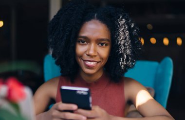 Siyah, neşeli, kıvırcık saçlı, günlük giysili kadın. Kafede sınav hazırlıkları sırasında akıllı telefon kullanarak sosyal medyayı kontrol ediyor.