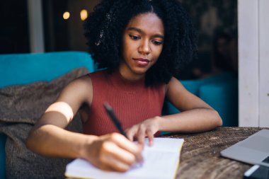 Kıvırcık saçlı, ciddi, siyah bir kadın dizüstü bilgisayarla çalışıyor. Not defterine not alıyor. Yazın kentteki kafede oturuyor.