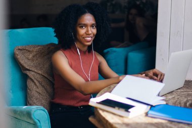 Kıvırcık saçlı, neşeli, etnik bir kadın. Yazın kafede elinde kahve not defteriyle dizüstü bilgisayarında yazı yazıyor.