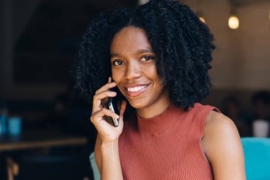 Neşeli kıvırcık saçlı, günlük giysiler içinde, cep telefonuyla konuşan, yazın açık terasta, mavi kanepede bir fincan kahve içen bir etnik kadın.