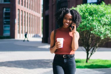 Mutlu etnik bayan, günlük giysiler içinde elinde kahve bardağıyla yazın şehirde cep telefonundan arama yapıyor.