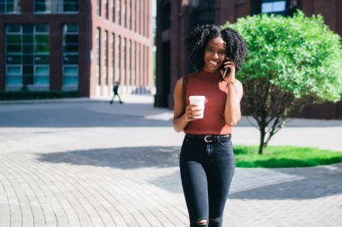 Mutlu etnik bayan, günlük giysiler içinde elinde kahve bardağıyla yazın şehirde cep telefonundan arama yapıyor.