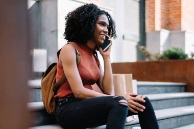 Sırt çantalı, sırt çantalı, cep telefonlu, gülen siyah kadın yazın granit merdivenlerde oturuyor.