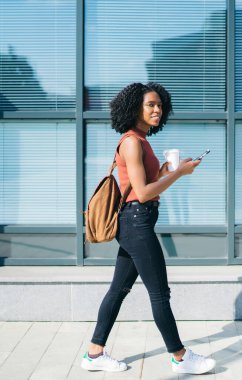 Sırt çantasındaki kahve fincanıyla şehirde yaz aylarında sokakta cep telefonundan mesajlaşan sıska, etnik bir kadın.