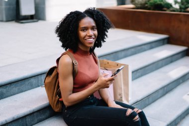 Sıradan giyinen, siyah kıvırcık saçlı kadın merdivenlerde oturup kameraya bakarak rahatlamak için akıllı telefon kullanıyor.