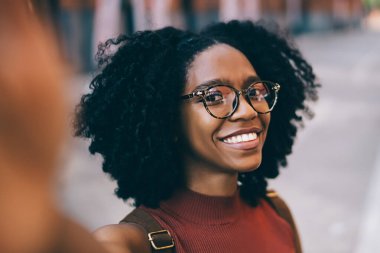 Genç, mutlu, gözlüklü, günlük kıyafetler giyen, şehirde selfie çekerken kameraya bakan Afro-Amerikan bir kadın.