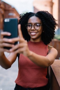 Afro saçlı, sıradan giyinmiş, gözlüklü, cep telefonu kullanan ünlü şehrin caddesinde selfie çeken mutlu bir zenci kadın.