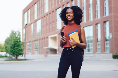 Mutlu Afro-Amerikan kıvırcık saçlı, kolsuz gömlekli, akıllı telefonlu, elinde kitaplarla yaz aylarında gezen kadın.