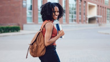Mutlu Afro-Amerikan kıvırcık saçlı, kolsuz tişörtlü, akıllı telefon kitaplarıyla yürüyen yaz aylarında el ele gezen bej sırt çantalı kadın.