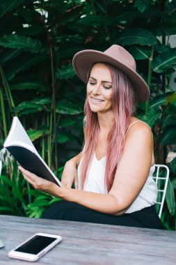 Şapkalı ve boyalı saçlarıyla genç ve çekici bir kadın. Tropik çalıların yanındaki masada oturuyor.