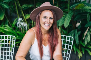 Masada oturmuş dışarıda akıllı telefon kullanırken kameraya bakan şapkalı, gülümseyen güzel bir kadın.