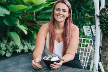 Dost canlısı, boyalı saçlı, gülümseyen ve kameraya bakan yetişkin bir kadın sabah tropikal bahçede kahvaltı ederken sağlıklı bir yemekle masada oturuyor.