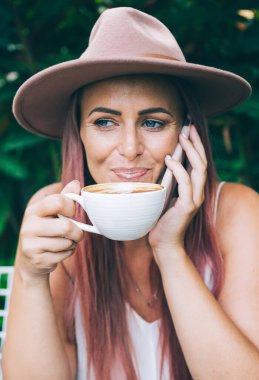 Neşeli gülümseyen, şapkalı, akıllı telefonlu ve kahvesini yudumlayan modern kadın tropikal yaz gününde açık terasta oturuyor.