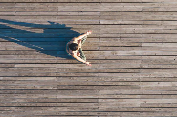 Yukarıdaki spor elbiseli kadın Lotus pozisyonunda otururken ahşap zeminde yoga yapıyor ve arkadan gölgesi düşüyor.