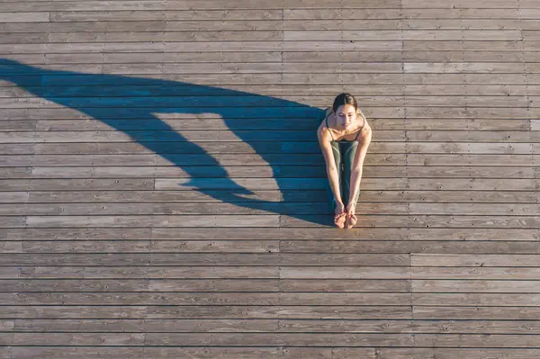 Tahta zeminde oturup kameraya bakarken, kollarını bacakları boyunca uzatan ve esneklik gösteren tanınmayacak kadar sağlıklı bir kadının üst görüntüsü.