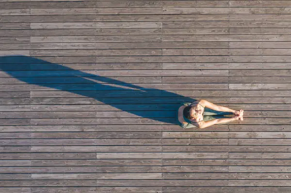 Spor giyim sektöründe aktif bir kadın, tahta zeminde oturur ve sabah antrenmanında kameraya bakarken esneme hareketleri yapar.