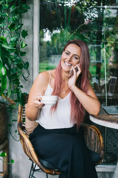 Yeşil bitkilerle çevrili açık kafede dizüstü bilgisayarı olan rahat, mutlu bir kadın elinde fincanla oturmuş akıllı telefonuyla konuşuyor.