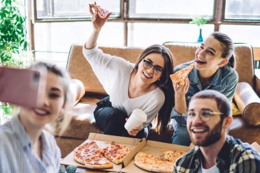 Bir grup neşeli genç, lezzetli pizzaları ve hazır içecekleriyle oturma odasındaki partide gülümseyip selfie çekiyorlar.