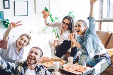 Dost canlısı gençler pizza yiyor ve cep telefonlarında selfie çekiyor. El kol hareketleri yapıyor ve iki parmağını kanepe ve yerde bacak bacak üstüne atmış bir şekilde gösteriyor.
