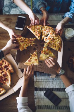 Yukarıdan gelen sabırsız arkadaşlar cep telefonlarını masaya koyuyor ve sebzeli ve sosisli pizza dilimleri yiyorlar.