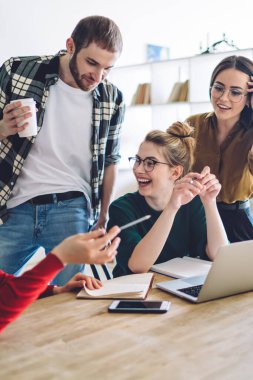 Modern apartmandaki rahat bir odada ev ödevi yaparken gülen bayan arkadaşlarının yanında duran içki ısmarlayan genç adam.