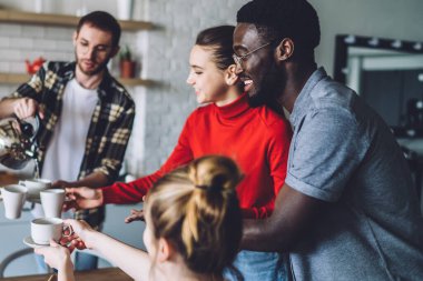 Modern apartmanın mutfağındaki mukavva kutularda servis edilmiş pizzayla masaya otururken mutlu insanlara çay dolduran bir adam.