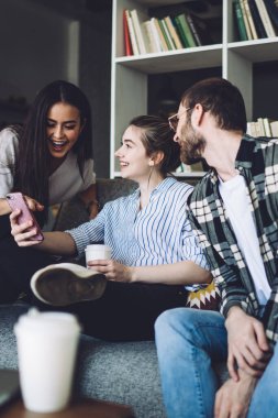 Heyecanlı koyu renk saçlı, bronz tenli kadın akıllı telefonu işaret ediyor ve çizgili gömlekli genç bayanla gülüyor ve gözlüklü ve kareli gömlekli adamla gülüyor.