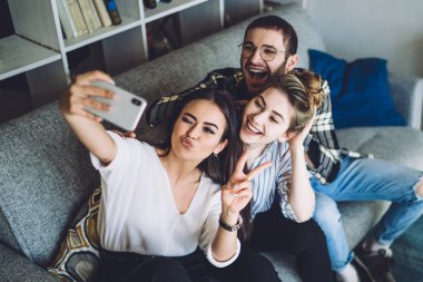Yukardan gelen koyu renk saçlı, etkileyici kadın mutlu bir kadınla selfie çekiyor ve modern bir dairede gözlüklü, neşeli bir adam.