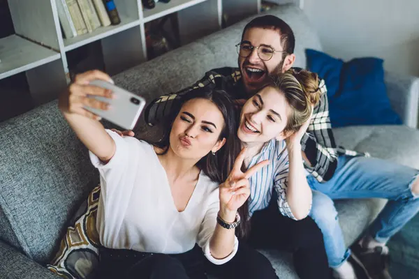 Yukardan gelen koyu renk saçlı, etkileyici kadın mutlu bir kadınla selfie çekiyor ve modern bir dairede gözlüklü, neşeli bir adam.