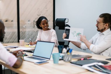 Modern ofis çalışma alanındaki dizüstü bilgisayarlarla dolu masada oturan meslektaşlarına entrika gösteren çok ırklı bir ekip çalışanı.