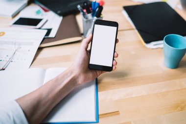 Yukarıdan tanınmayan bir adamın ofisinde kırtasiyeyle çalışırken boş beyaz ekranlı modern akıllı telefonu kullanması.