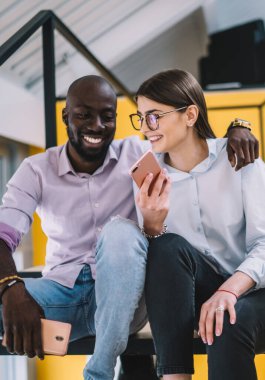 Genç bir kadın, birlikte sanat alanında oturup sosyal medyadan komik resimlere gülerken neşeli Afrikalı Amerikalı bir erkeğe cep telefonu gösteriyor.