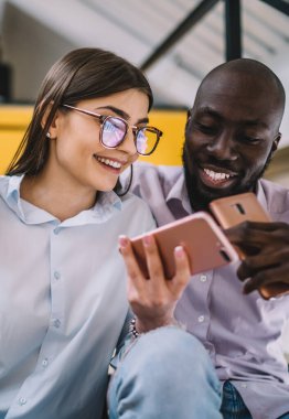 Genç Afro-Amerikan erkek, neşeli kız arkadaşını kucaklıyor ve internette komik fotoğraflar çekerken birlikte oturuyor ve cep telefonu kullanıyor.