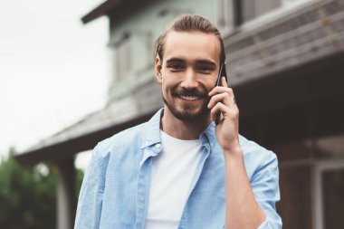 Günlük kıyafetli neşeli genç adam banliyö evinin bahçesinde dikilirken kameraya bakıyor ve telefona cevap veriyor.