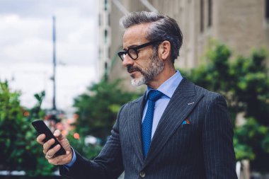 Olgun yakışıklı, meşgul, gözlüklü, gri saçlı, resmi takım elbiseli, internette sörf yapan ve modern dijital akıllı telefondan kentsel arka planda çalışan biri.