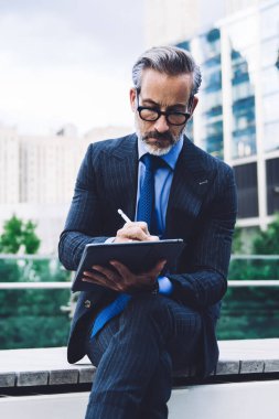 New York binalarının arka planında otururken Stylus 'la tablet kullanan konsantre orta yaşlı takım elbiseli ve gözlüklü bir adam.