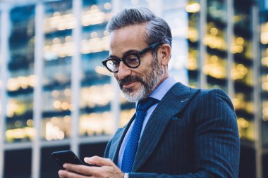 Yakışıklı, gri saçlı, son moda saç kesimli, siyah ceketli, mavi kravatlı ve New York 'ta bulanık arka planda cam çitle akıllı telefondan mesaj atan bir iş adamı.