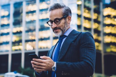 Yakışıklı, gri saçlı, son moda saç kesimli, siyah ceketli, mavi kravatlı ve New York 'ta bulanık arka planda cam çitle akıllı telefondan mesaj atan bir iş adamı.