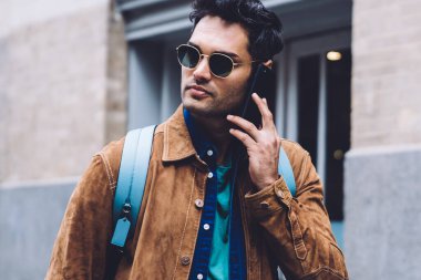 Gündelik giysiler ve güneş gözlüğü takmış, modern dijital akıllı telefondan arayan ve şehirde cadde boyunca yürürken gözlerini kaçıran genç, şık, ciddi bir erkek.