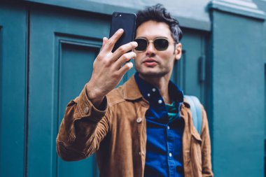 Sırt çantası ve güneş gözlüğü takmış yakışıklı genç adam New York caddesinin mavi binasında dikilirken selfie çekiyor.