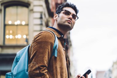 Gözlüklü, kahverengi süet ceketli, sırt çantalı ve kulaklıklı şık İspanyol bir erkek New York 'ta yaya geçidinde dikilip gözlerini kaçırıyor.