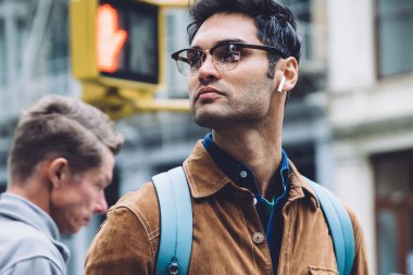 Gözlüklü, kahverengi süet ceketli, sırt çantalı ve kulaklıklı düşünceli bir İspanyol erkek New York caddesinde yeşil ışık beklerken müzik dinliyor ve başka tarafa bakıyor.