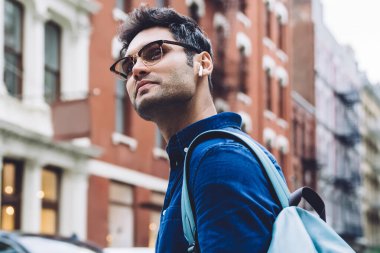 Gözlüklü, sırt çantalı ve akıllı telefonlu genç bir adamın kalabalık şehir caddesinde yürürken etrafa bakınışının yan görüntüsü