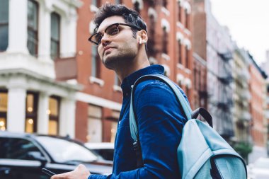 Gözlüklü, sırt çantalı ve akıllı telefonlu genç bir adamın kalabalık şehir caddesinde yürürken etrafa bakınışının yan görüntüsü