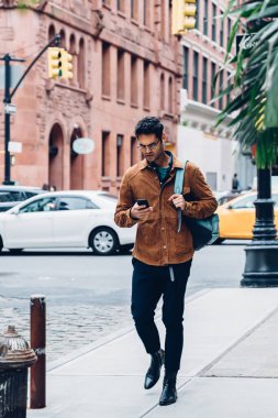 Gözlüklü ve ceketli havalı yetişkin bir adam. Sırt çantası taşıyor ve gün ışığında yaya geçidinde yürürken akıllı telefon kullanıyor.