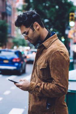 Kendine güvenen yakışıklı, ceketli ve gözlüklü yetişkin bir adamın akıllı telefon kullanarak kentsel yaya geçidinde durmasının yan görüntüsü.
