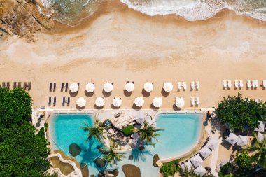 Açık havuzu ve güneş pansiyonları olan ilk hat plaj otelinin havadan görünüşü. Yaz tatillerinin kuş bakışı manzarası, turkuaz deniz suyu ve temiz kum kıyısındaki şemsiyeler.